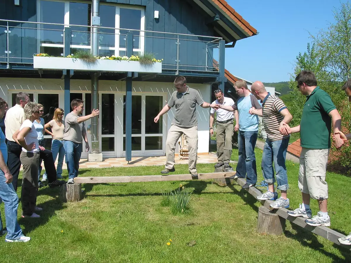 Eine Gruppe von Menschen steht gemeinsam auf einem schmalen Holzbalken in etwa 30 Zentimeter Höhe vom Boden. Die Teilnehmenden balancieren und halten sich an den Händen.