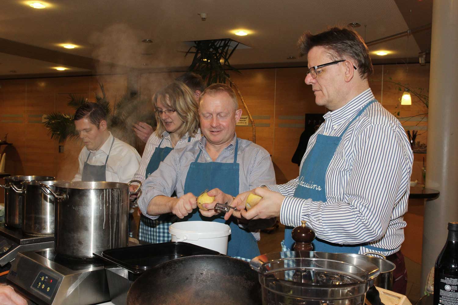 Eine Gruppe Menschen steht vor einigen Töpfen auf Herdplatten, zwei Männer schälen Kartoffeln. Teamkochen im Landhotel am Rothenberg