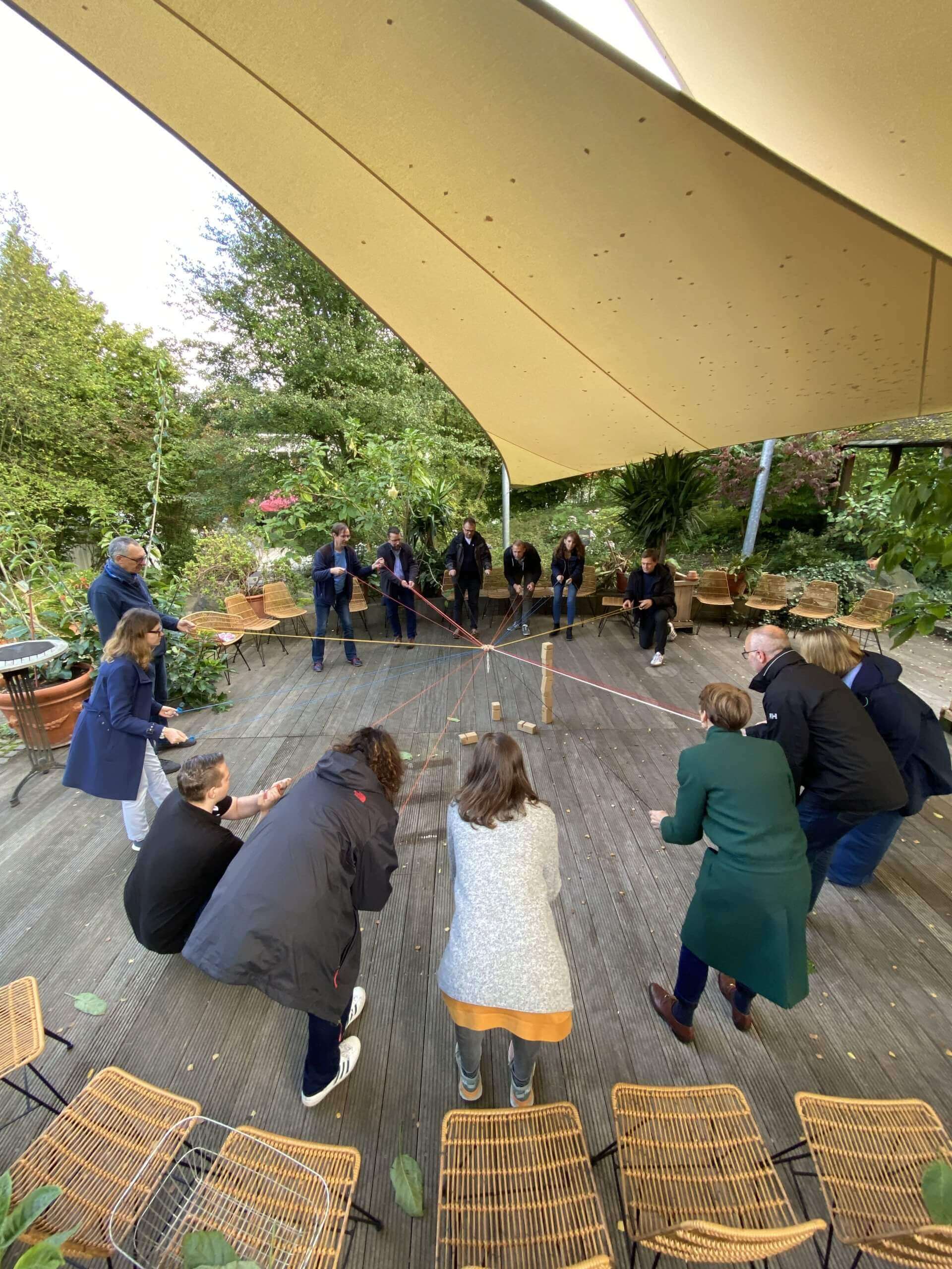 Gruppe von Menschen im Kreis stehend. In der Mitte des Kreises stehen Bausteine, die über ein Seil mit jedem Mensch verbunden sind.